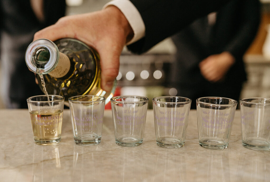 A hand in formal attire pours a golden beverage into shot glasses lined up on a marble counter, setting the celebratory mood at a northern California private estate wedding.