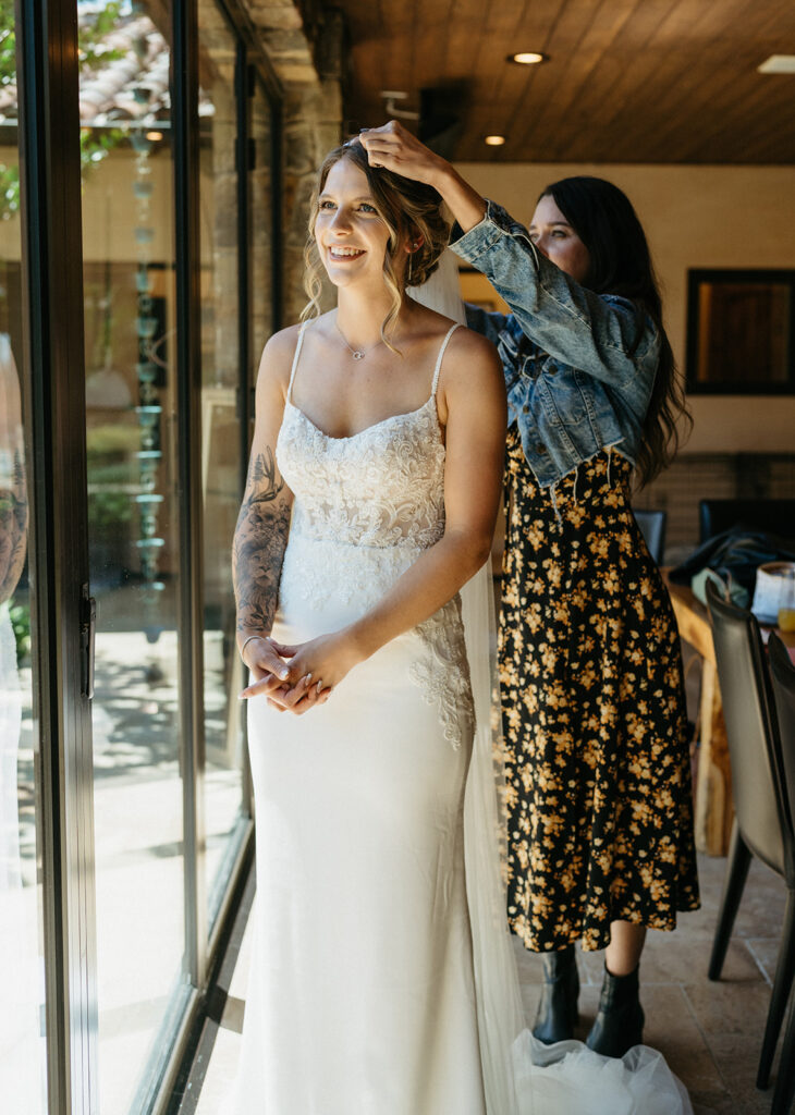 "A radiant bride in a detailed wedding gown stands by a window, getting a final hair touch-up from a friend dressed in a floral skirt and denim jacket at a northern California private estate wedding.