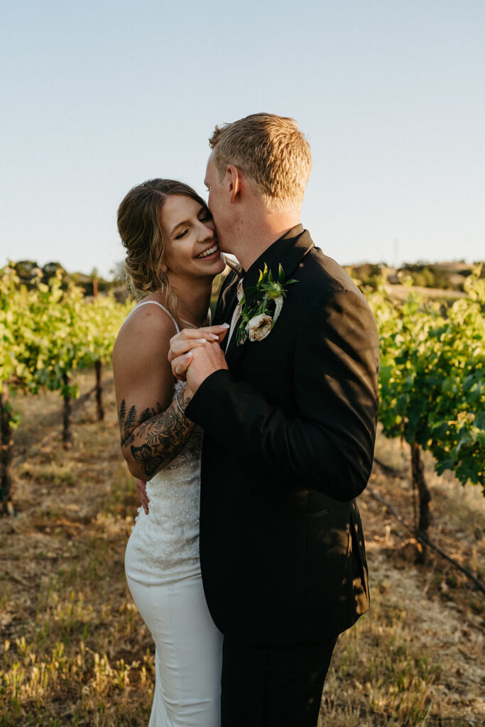 A couple embracing after their Northern California private estate wedding in a vineyard during golden hour, with the woman smiling and the man's face close to hers. The woman has a tattoo on her arm and wears a white dress, while the man is in a black suit with a boutonnière. 
