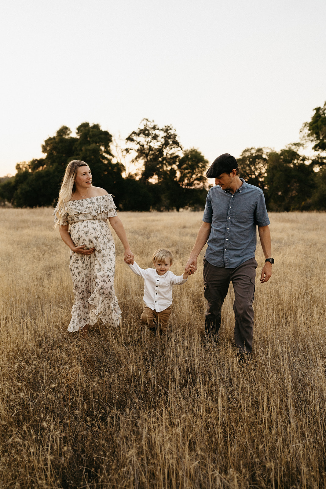 A family of three - soon to be four - walking in a field during golden hour.