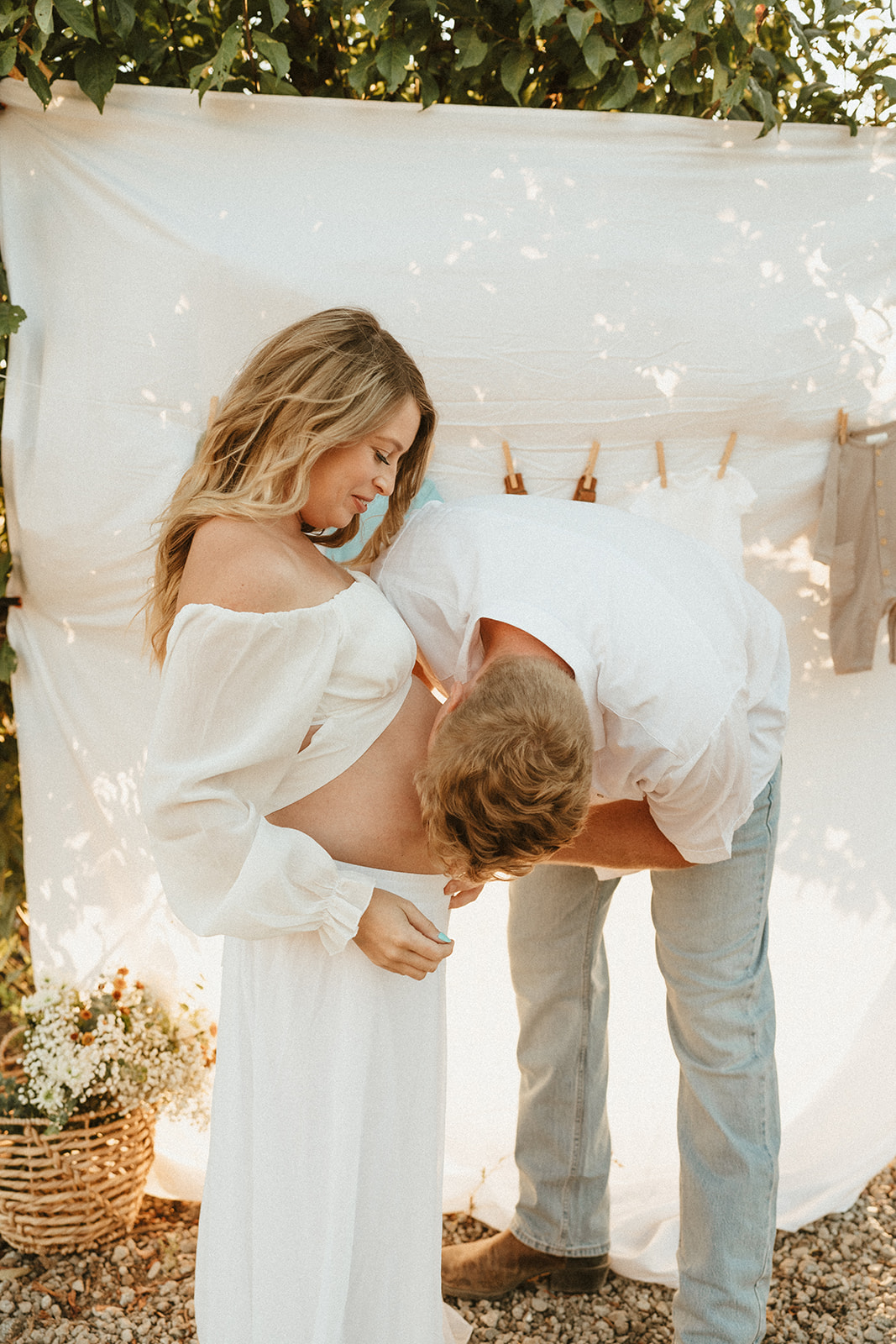 Blonde mother-to-be, radiantly pregnant with a white boho, bell-sleeved top and flowing white skirt stands as father-to-be kisses her belly. Pregnant mother and father stand in front of a flowing white sheet billowing in the wind. A clothesline lined with baby clothes is draped across the white sheet. The image is tinted with golden light from sunset. A basket of florals sits in the bottom left corner of the image.