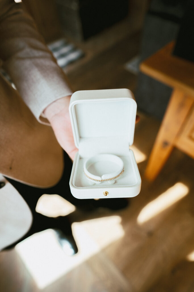 Groom holding a white box with a delicate wedding bracelet, preparing for his Tahoe Mountain Club ceremony in a rustic mountain lodge.
