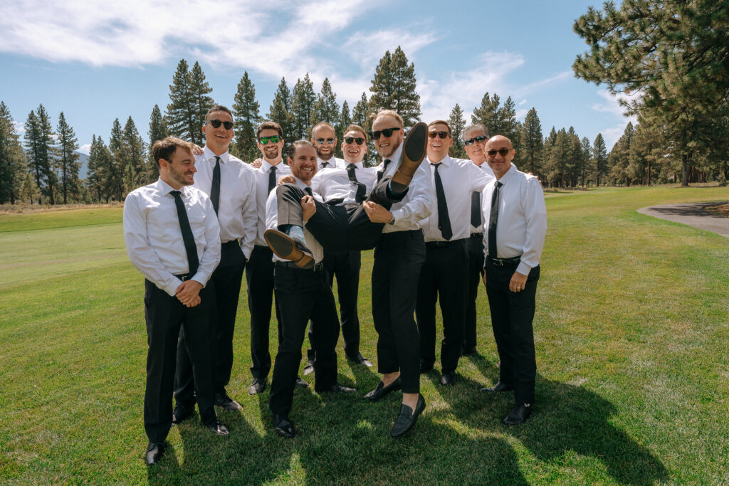 Groomsmen hoisting the groom in front of scenic pines, sharing a playful moment before the Tahoe Mountain Club ceremony.