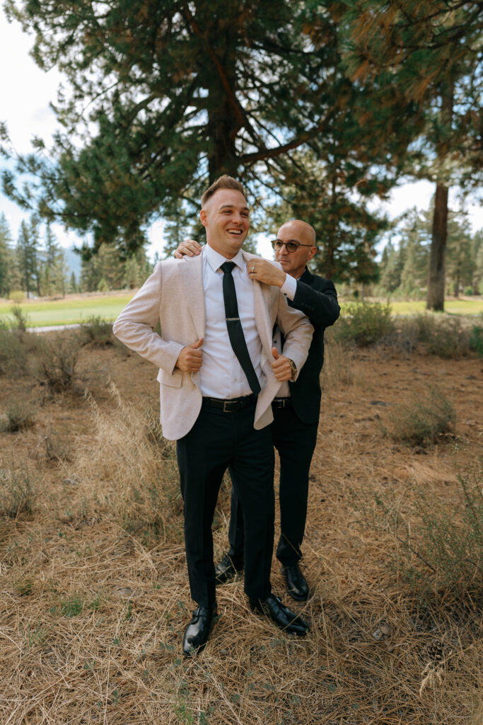 Groom wearing a beige blazer, smiling while a groomsman adjusts his jacket under pine trees at a Tahoe Mountain Club wedding.