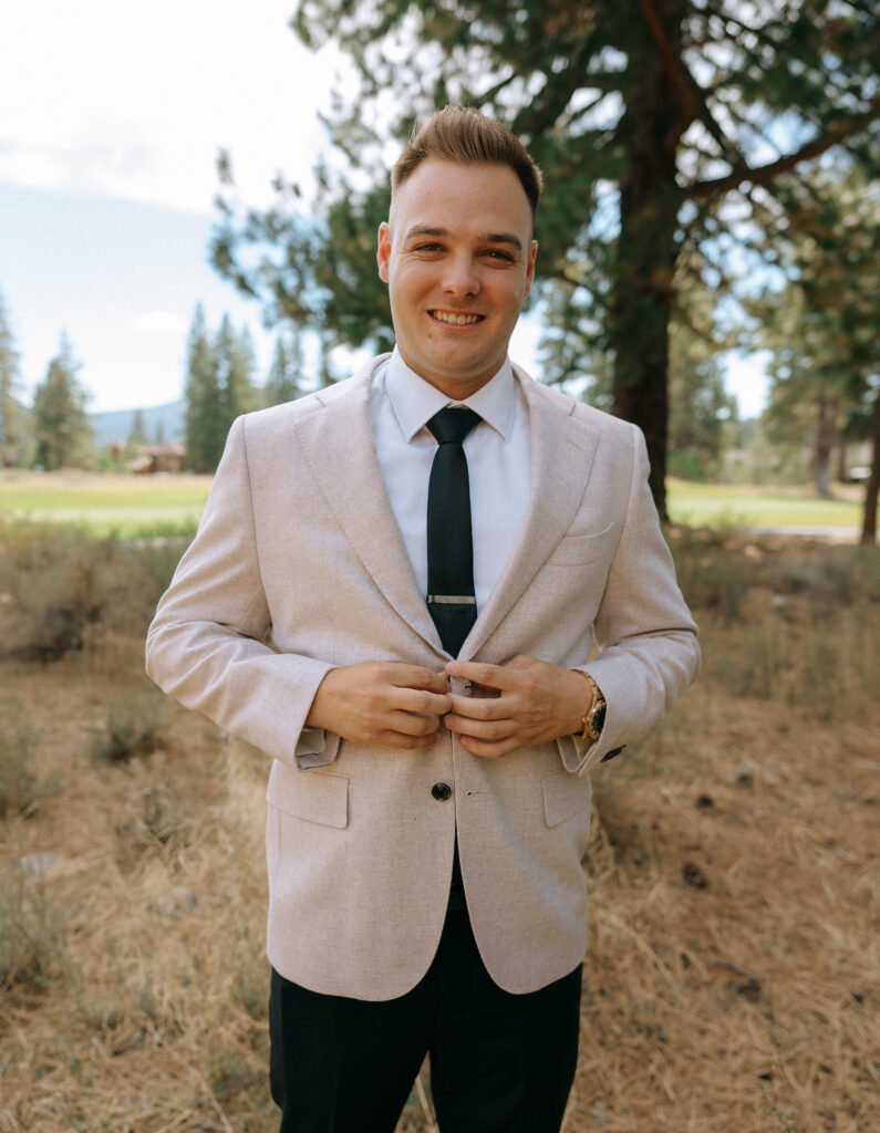 Groom adjusting his beige blazer among towering pines in Truckee, preparing for a Tahoe Mountain Club wedding in the Sierra Nevada.