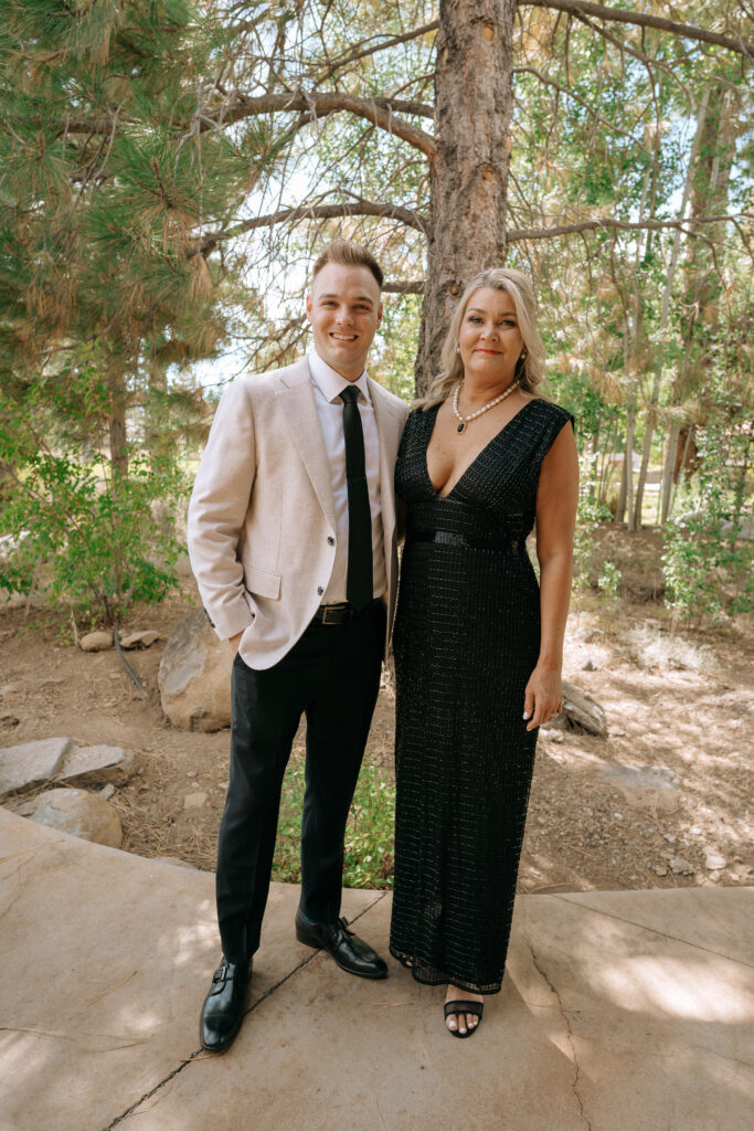 Groom in a beige blazer standing with a woman in a black dress under pine trees, moments before a Tahoe Mountain Club wedding in Truckee.