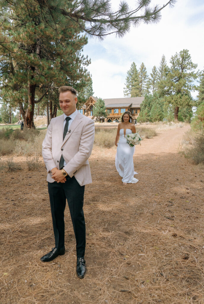 Bride approaches the groom from behind, holding her white bouquet in a rustic pine setting for their first look at Tahoe Mountain Club.