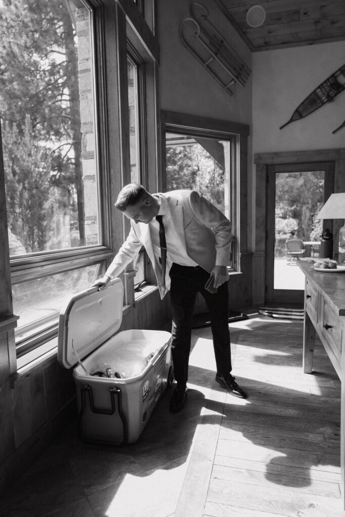 Groom in black-and-white, checking a cooler inside a sunny lodge, getting ready for his Tahoe Mountain Club wedding.