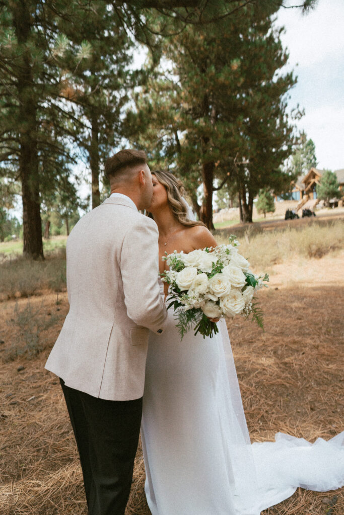 Bride and groom sharing a kiss in a pine forest near Tahoe Mountain Club, featuring a lush white rose bouquet and serene mountain wedding backdrop.