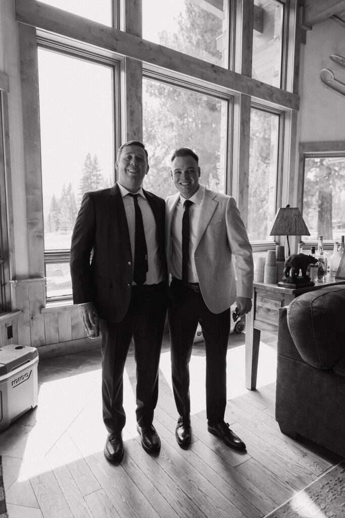 Groom and friend in black-and-white, standing by floor-to-ceiling windows in a rustic lodge, preparing for a Tahoe Mountain Club wedding.