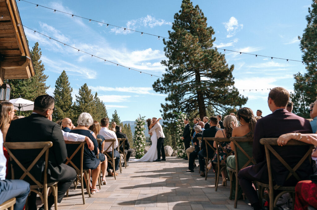 Bride and groom exchanging vows outdoors at Tahoe Mountain Club terrace ceremony with panoramic Lake Tahoe views, string lights, and elegant floral arch.