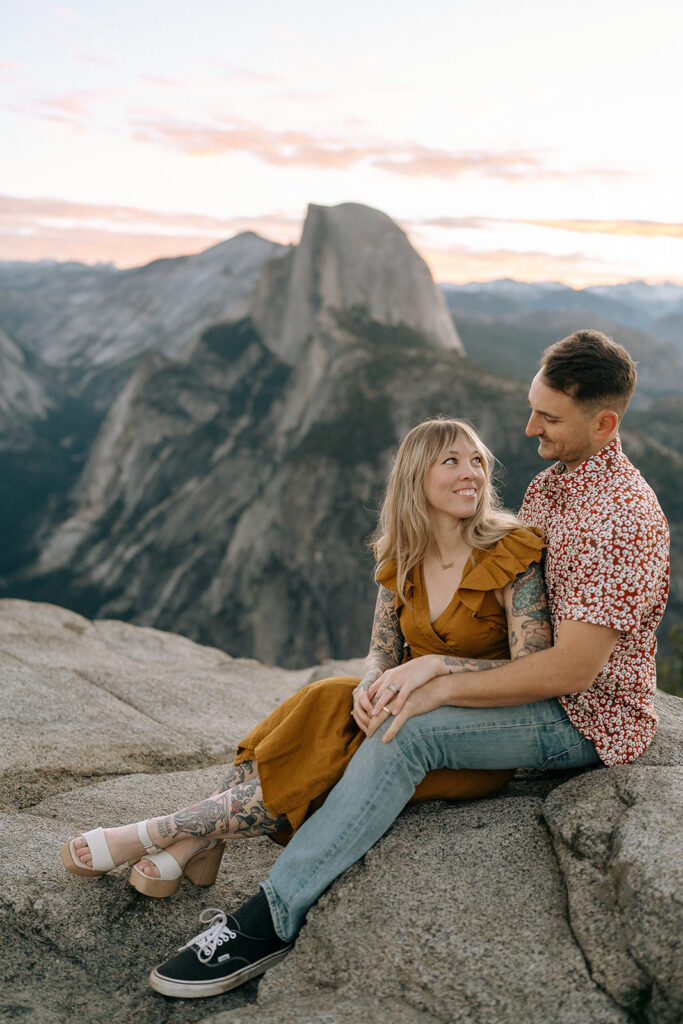 A couple sits together on a rocky ledge at Glacier Point overlooking Half Dome at sunset, documented by a Yosemite photographer.