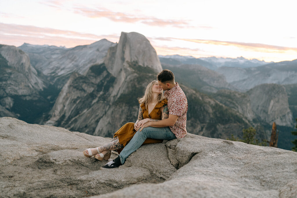 A couple sits side by side on a rocky ledge at Glacier Point with Half Dome in the distance, photographed by a Yosemite photographer at sunset.