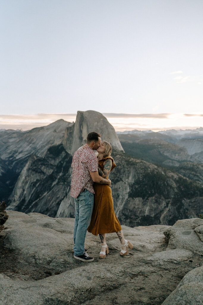 A couple stands together on a granite overlook with Half Dome rising in the background, captured by a Yosemite photographer.