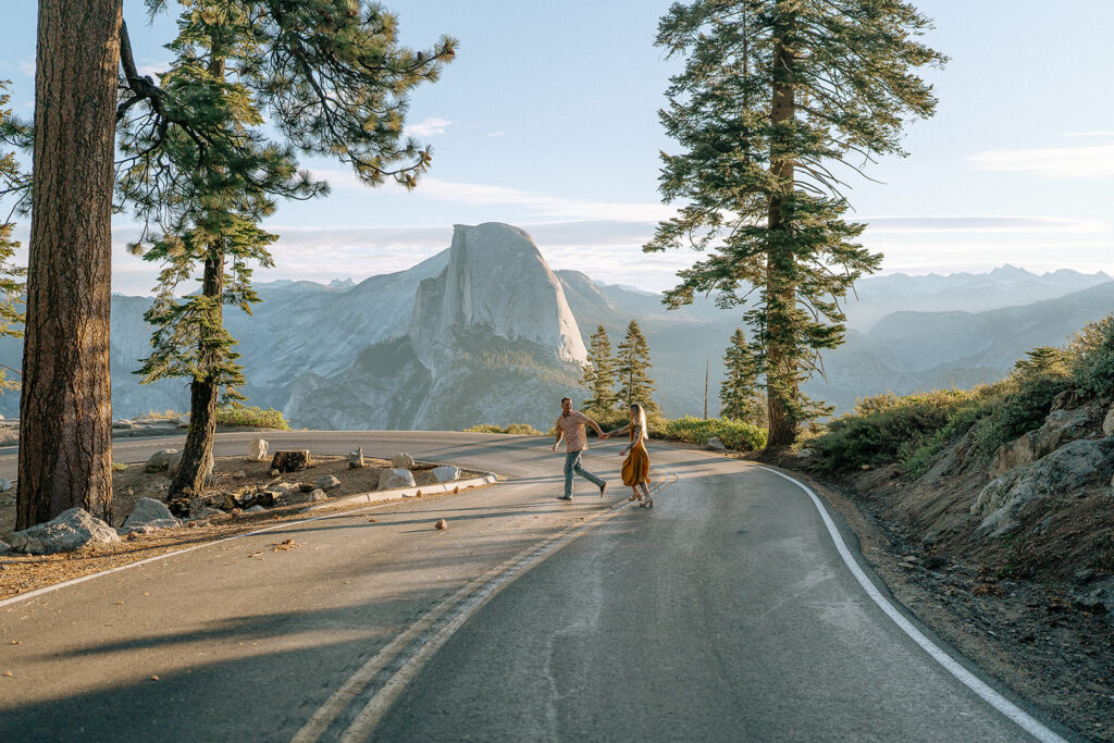 A couple playfully runs along Glacier Point Road with Half Dome in the background, all photographed by a Yosemite photographer.