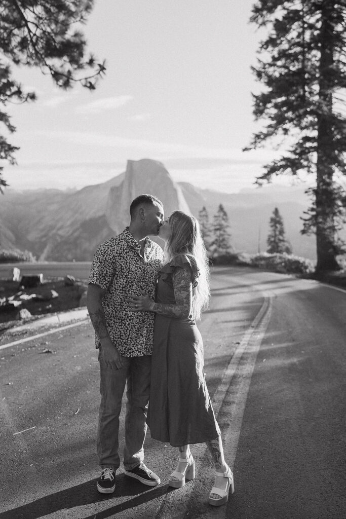 A black and white photo of a couple leaning in for a kiss on Glacier Point Road, featuring a distant Half Dome, taken by a Yosemite photographer.