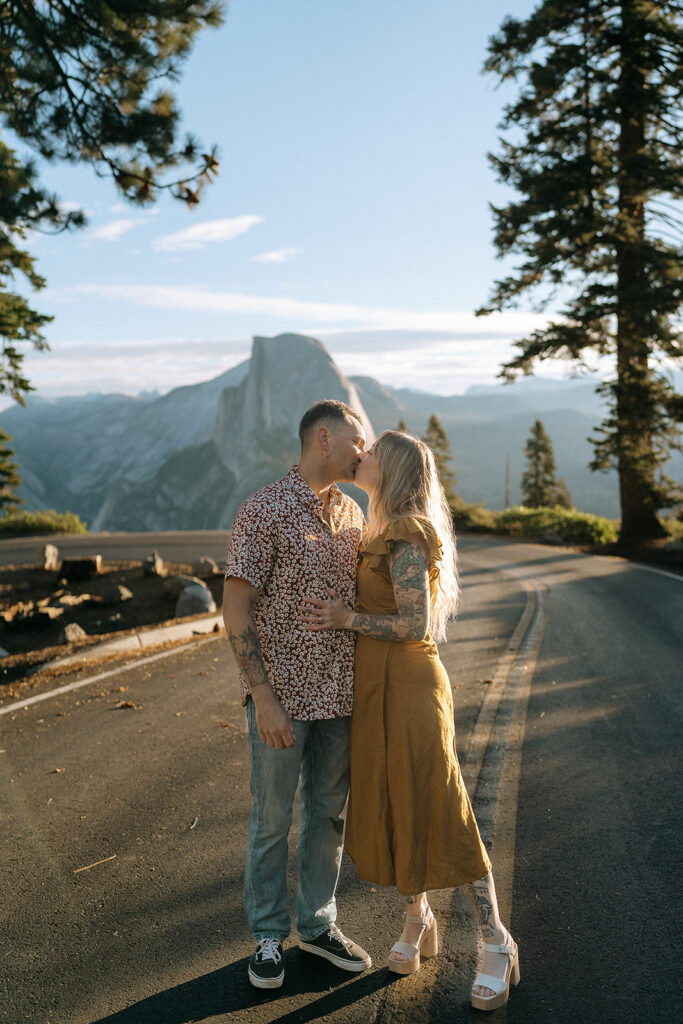 A couple shares a kiss on Glacier Point Road, framed by towering pine trees and Half Dome, captured by a Yosemite photographer.